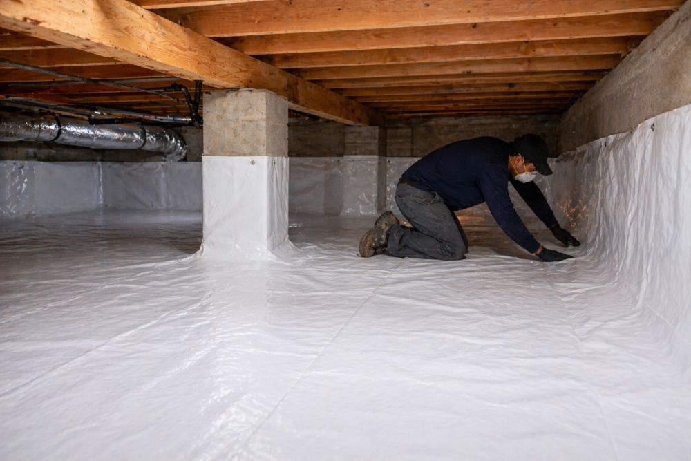 This image shows vapor barrier installation in Burbank, CA by Pure Eco Inc inside a residential crawl space. A technician installs a durable plastic vapor barrier across the soil floor and along the lower foundation walls to control moisture and reduce humidity buildup. The crawl space includes exposed wooden beams and structural supports with the barrier laid flat and secured for full coverage. This process helps prevent mold growth, protect insulation, and improve indoor air quality. Pure Eco Inc provides crawl space and moisture control services across Burbank. Homeowners can call (877) 870-7998 to schedule an inspection and installation service.