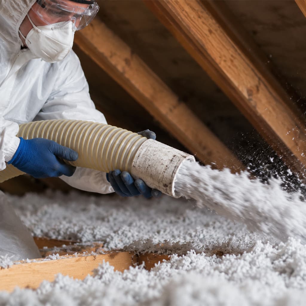 Close-up of a technician installing blown-in insulation in an attic using a hose while wearing protective gear.