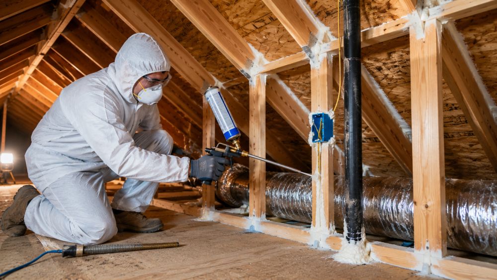 Pure Eco performs professional air sealing in Sherman Oaks, CA to reduce energy loss and improve indoor comfort. This image shows a trained technician applying expanding foam sealant around framing joints, utility penetrations, and attic openings inside a residential attic. Visible attic components include wooden rafters, roof decking, electrical boxes, and HVAC ductwork that require proper sealing to reduce conditioned air loss. Pure Eco, located in Los Angeles, California, provides air sealing, insulation, and energy efficiency services throughout Sherman Oaks and surrounding areas.