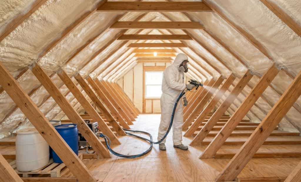 A professional insulation technician in a protective suit spraying expanding foam insulation between the wooden roof rafters of an attic.